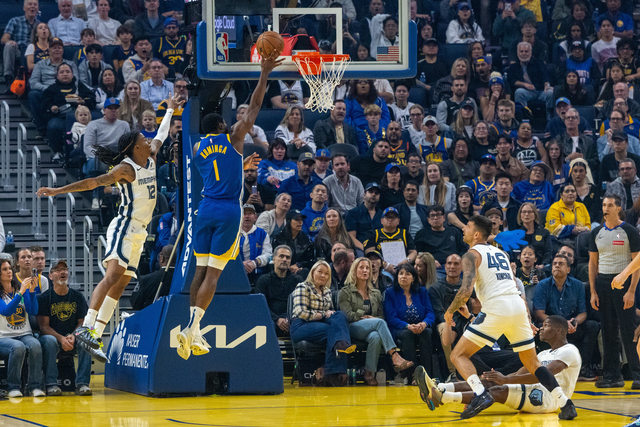 A basketball player in blue jumps to dunk the ball while a defender in white tries to block him; a fallen player and a referee watch nearby.